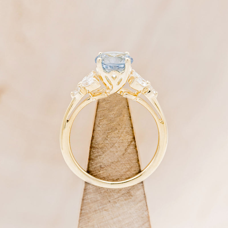 Shown here is "Blossom", a round cut aquamarine women's engagement ring with leaf-shaped diamond accents, side view on stand. Many other center stone options are available upon request.