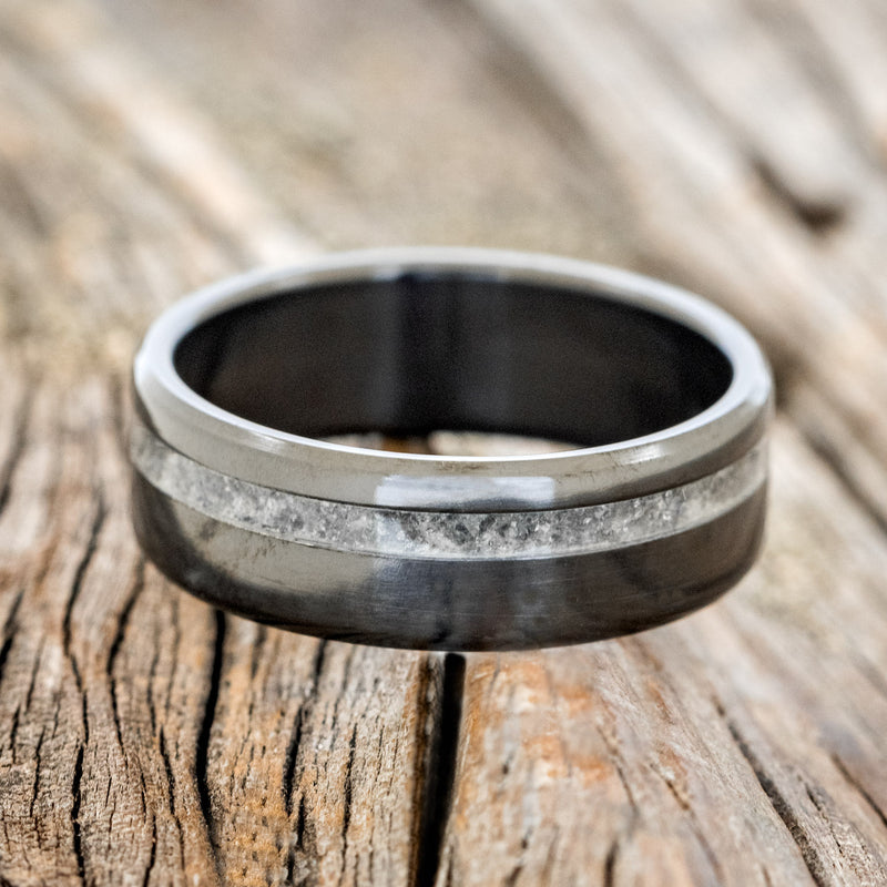 Shown here is "Vertigo", a custom, handcrafted men's wedding ring featuring an offset moonstone inlay on a fire-treated black zirconium band, laying flat. Additional inlay options are available upon request.