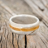 Shown here is "Asher", a custom, handcrafted men's wedding ring featuring a Bethlehem olive wood and a 14K yellow gold inlay on a Damascus steel band, laying flat. Additional inlay options are available upon request.