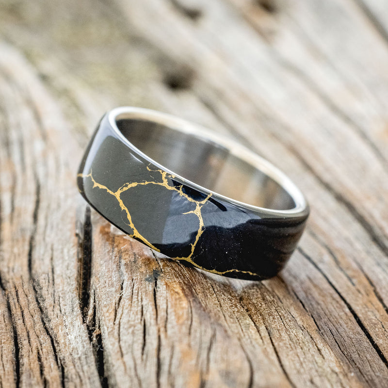 Shown here is "Haven", a custom, handcrafted men's wedding ring featuring a black and gold TruStone overlay on a titanium band, tilted left. Additional overlay options are available upon request.
