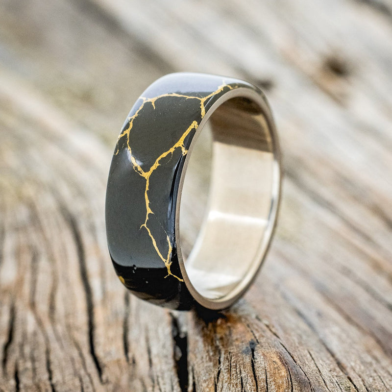 Shown here is "Haven", a custom, handcrafted men's wedding ring featuring a black and gold TruStone overlay on a titanium band, upright facing left. Additional overlay options are available upon request.