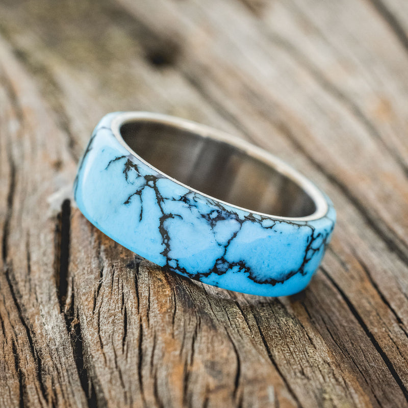Shown here is "Haven", a custom, handcrafted men's wedding ring featuring a turquoise and black matrix TruStone overlay on a titanium band, tilted left. Additional overlay options are available upon request.