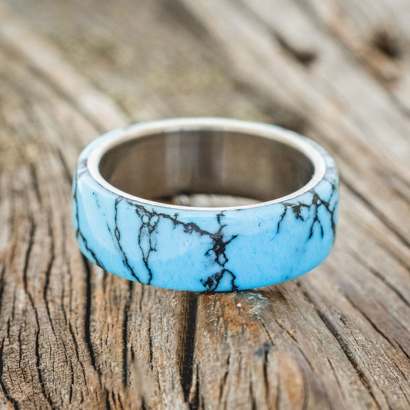 Shown here is "Haven", a custom, handcrafted men's wedding ring featuring a turquoise and black matrix TruStone overlay on a titanium band, laying flat. Additional overlay options are available upon request.