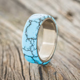 Shown here is "Haven", a custom, handcrafted men's wedding ring featuring a turquoise and black matrix TruStone overlay on a titanium band, upright facing left. Additional overlay options are available upon request.