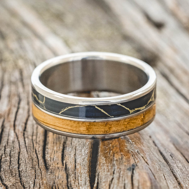 Shown here is "Dyad", a custom, handcrafted men's wedding ring featuring 2 channels with whiskey barrel oak and black and gold matrix TruStone inlays, laying flat. Additional inlay options are available upon request.