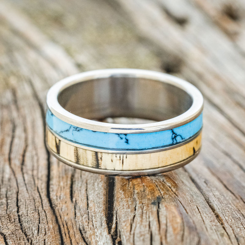 Shown here is "Dyad", a custom, handcrafted men's wedding ring featuring 2 channels with spalted maple wood and turquoise with black matrix TruStone inlays, laying flat. Additional inlay options are available upon request.