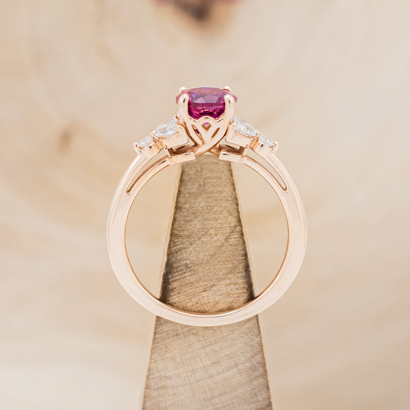  Shown here is "Blossom", a round cut lab-created ruby women's engagement ring with leaf-shaped diamond accents, side view on stand. Many other center stone options are available upon request.