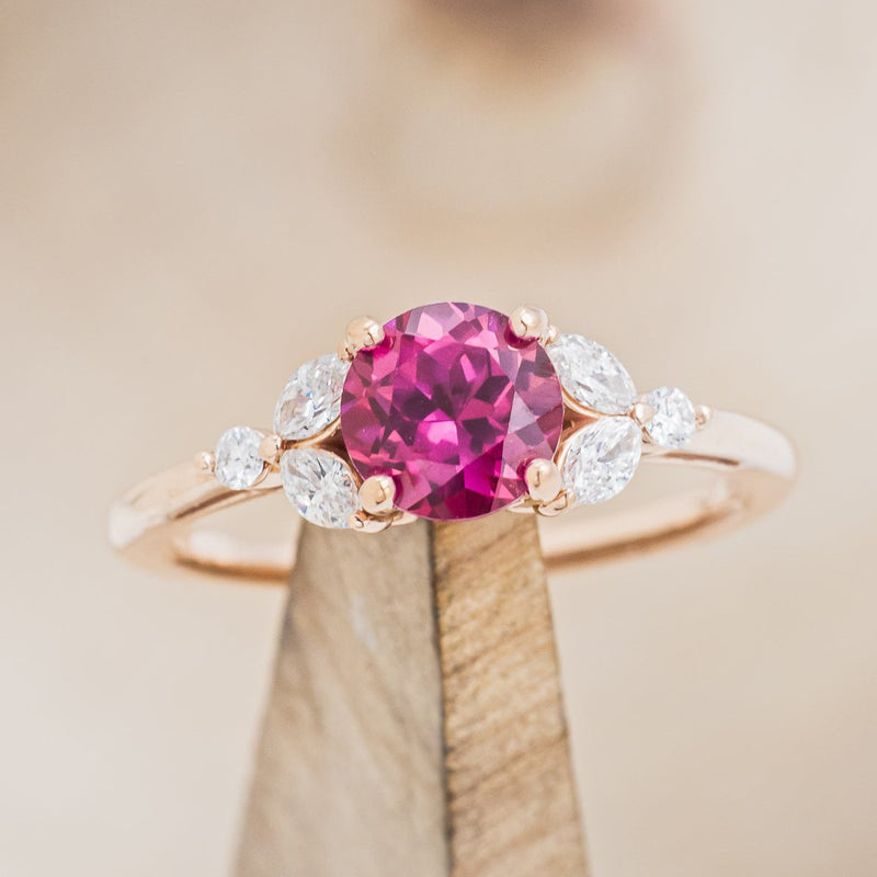 Shown here is "Blossom", a round cut lab-created ruby women's engagement ring with leaf-shaped diamond accents, on stand front facing. Many other center stone options are available upon request. 