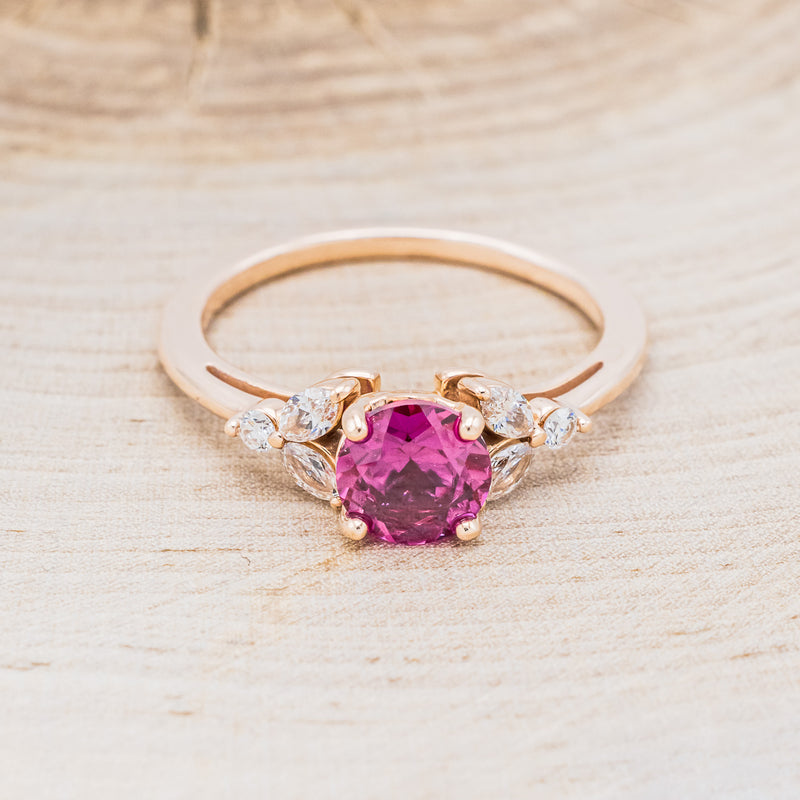 Shown here is "Blossom", a round cut lab-created ruby women's engagement ring with leaf-shaped diamond accents, front facing. Many other center stone options are available upon request.