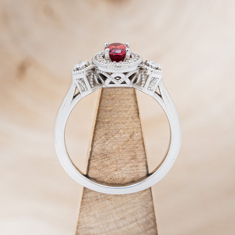 Shown here is "Amelia", a vintage-style lab-created ruby women's engagement ring with a diamond halo and diamond accents, side view on stand. Many other center stone options are available upon request.