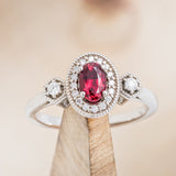 Shown here is "Amelia", a vintage-style lab-created ruby women's engagement ring with a diamond halo and diamond accents, on stand front facing. Many other center stone options are available upon request. 