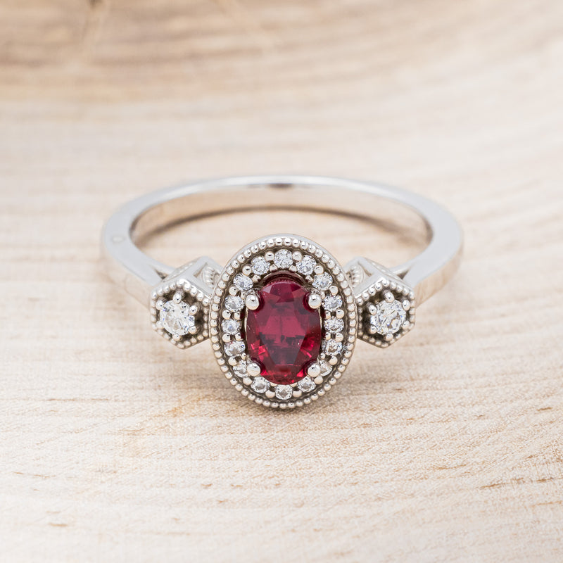 Shown here is "Amelia", a vintage-style lab-created ruby women's engagement ring with a diamond halo and diamond accents, front facing. Many other center stone options are available upon request.