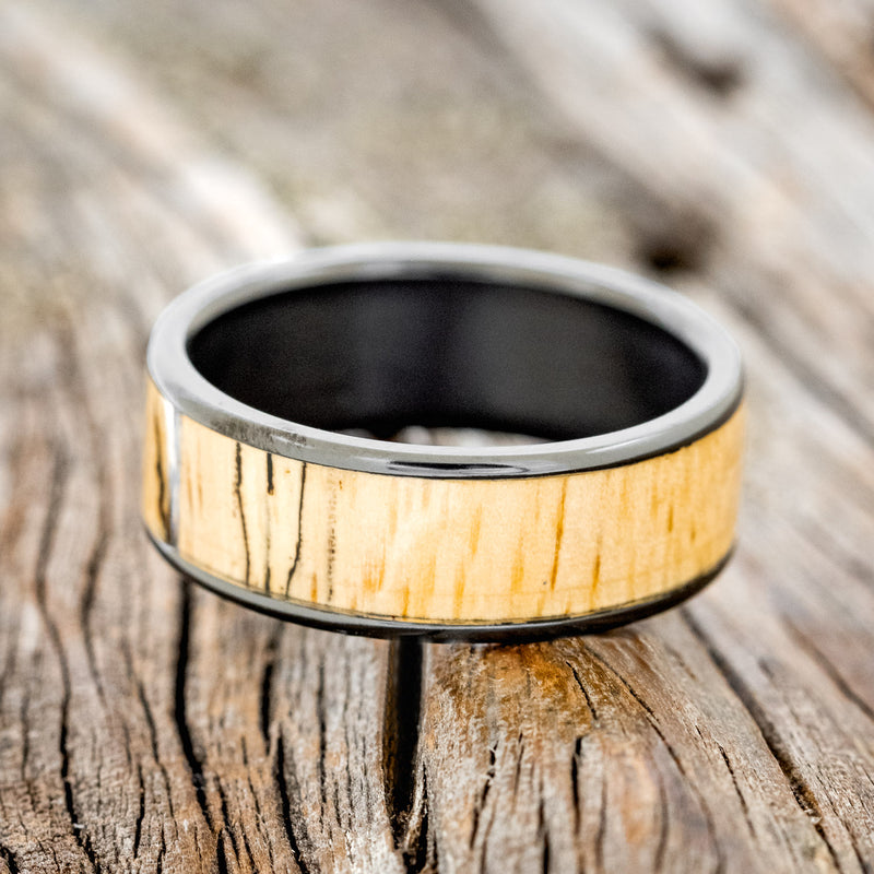 Shown here is "Rainier", a custom, handcrafted men's wedding ring featuring spalted maple wood inlay, shown here on a fire-treated black zirconium band, laying flat. Additional inlay options are available upon request.