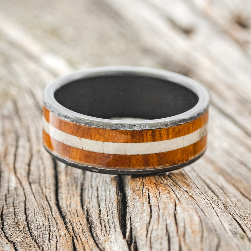Shown here is "Rainier", a custom, handcrafted men's wedding ring featuring an ironwood and antler inlay on a hammered, fire-treated black zirconium band, laying flat. Additional inlay options are available upon request.