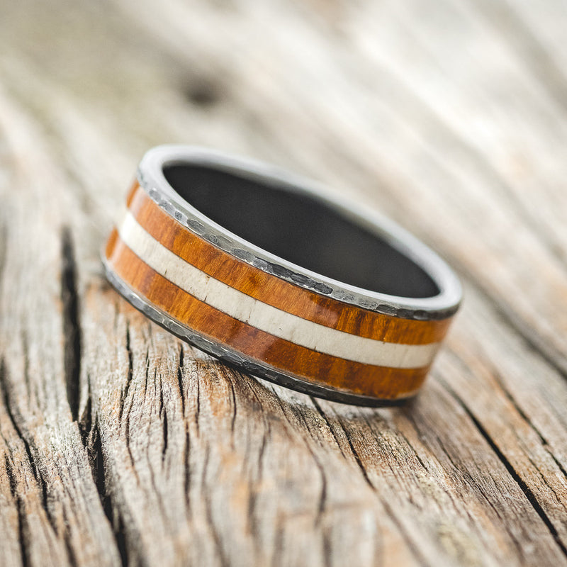 Shown here is "Rainier", a custom, handcrafted men's wedding ring featuring an ironwood and antler inlay on a hammered, fire-treated black zirconium band, tilted left. Additional inlay options are available upon request.
