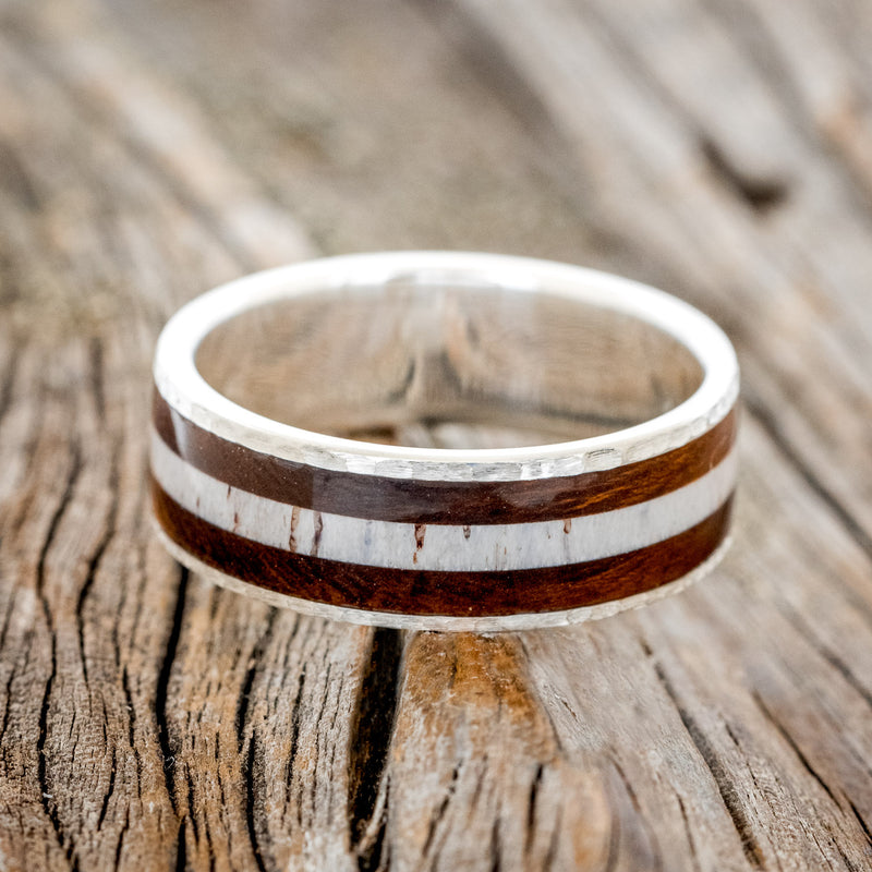 Shown here is "Rainier", a custom, handcrafted men's wedding ring featuring an ironwood and antler inlay on a hammered band, laying flat. Additional inlay options are available upon request.