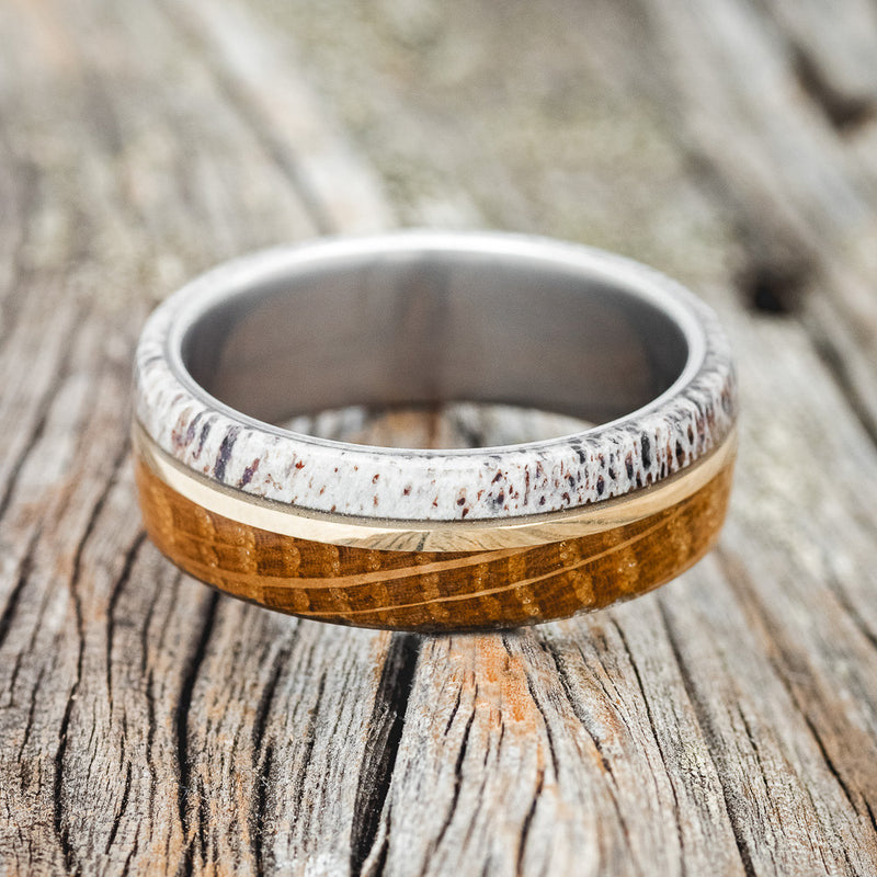 Shown here is "Golden", a handcrafted men's wedding ring featuring a whiskey barrel and antler overlay with a 14K yellow gold inlay, laying flat. Additional inlay options are available upon request.
