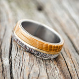 Shown here is "Golden", a handcrafted men's wedding ring featuring a whiskey barrel and antler overlay with a 14K yellow gold inlay, tilted left. Additional inlay options are available upon request.