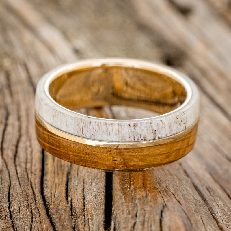 Shown here is "Golden", a handcrafted men's wedding ring featuring a whiskey barrel and antler overlay with a 14K yellow gold inlay, laying flat. Additional inlay options are available upon request.