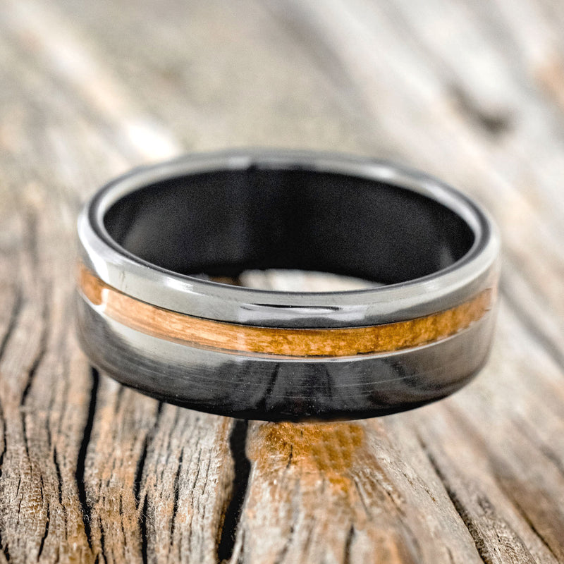 Shown here is "Vertigo", a custom, handcrafted men's wedding ring featuring a whiskey barrel inlay, shown here on a fire-treated black zirconium band, laying flat. Additional inlay options are available upon request.