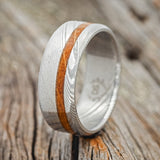 Shown here is "Vertigo", a custom, handcrafted men's wedding ring featuring a whiskey barrel inlay, shown here on a Damascus steel band, upright facing left. Additional inlay options are available upon request.