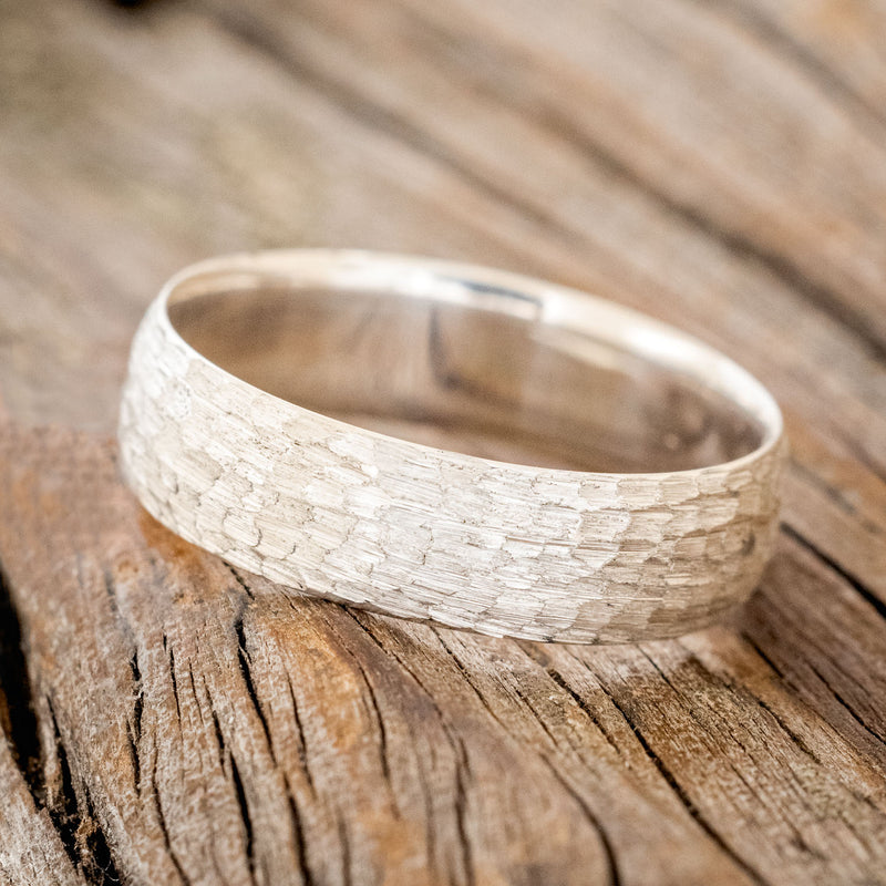 Shown here is a handcrafted men's wedding ring featuring a solid band with a domed profile and hammered finish, tilted left. Additional inlay options are available upon request.