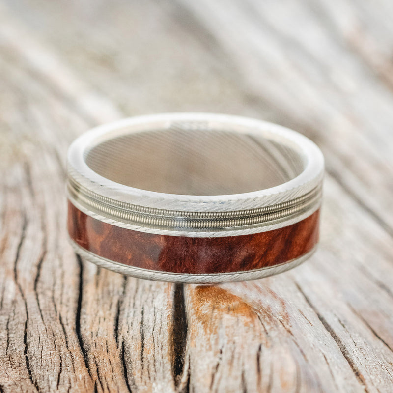 Shown here is "Raptor", a custom, handcrafted men's wedding ring featuring 2 channels with guitar string and redwood inlays, laying flat. Additional inlay options are available upon request.