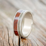 Shown here is "Raptor", a custom, handcrafted men's wedding ring featuring 2 channels with guitar string and redwood inlays, upright facing left. Additional inlay options are available upon request.