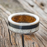 Shown here is "Flynn", a custom, handcrafted men's wedding ring featuring elk tooth & charred whiskey barrel oak inlays with a whiskey barrel lining, laying flat. Additional inlay options are available upon request.