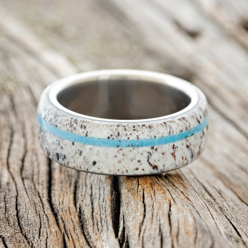 Shown here is "Remmy", a custom, handcrafted men's wedding ring featuring an antler overlay with a turquoise inlay, laying flat. Additional inlay options are available upon request.