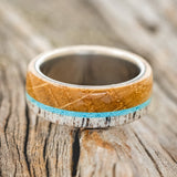 Shown here is "Banner", a custom, handcrafted men's wedding ring featuring whiskey barrel oak and antler overlay with a turquoise inlay on a titanium band, laying flat. Additional inlay options are available upon request.