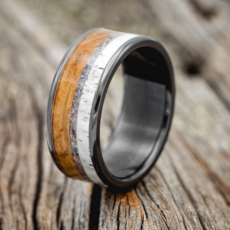 Shown here is "Rainier", a custom, handcrafted men's wedding ring featuring whiskey barrel oak, crushed charoite stone, and elk antler, shown here on a fire-treated black zirconium band, upright facing left. Additional inlay options are available upon request.