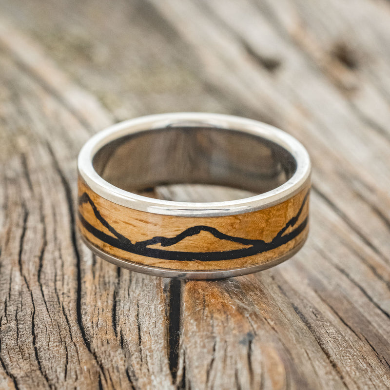 Shown here is "The Expedition", a custom, handcrafted men's wedding ring featuring a mountain engraving with whiskey barrel oak and charred whiskey barrel inlays, laying flat. Additional inlay options are available upon request.
