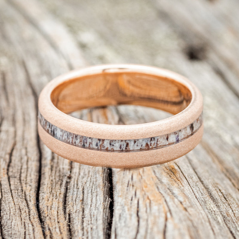 Shown here is "Vertigo", a custom, handcrafted men's wedding ring featuring an offset antler inlay on a sandblasted 14K gold band, laying flat. Additional inlay options are available upon request.