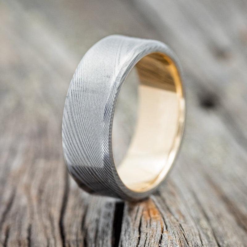 Shown here is "Vulcan", a handcrafted men's wedding ring featuring a hand-forged stainless Damascus steel overlay on a 14K gold band, upright facing left. Additional inlay options are available upon request.