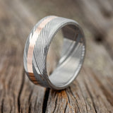 Shown here is "Vertigo", a custom, handcrafted men's wedding ring featuring a 14K rose gold inlay, shown here on a Damascus steel band, upright facing left. Additional inlay options are available upon request.