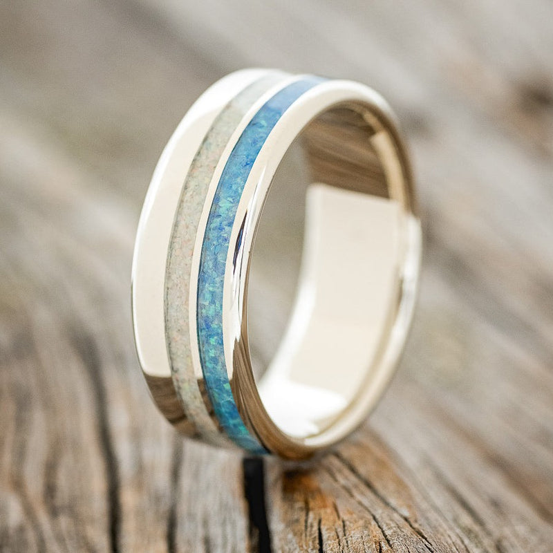 Shown here is "Cosmo", a custom, handcrafted men's wedding ring featuring two offset inlays with blue opal and fire and ice opal on a 14K gold band, upright facing left. Additional inlay options are available upon request.