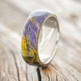 Shown here is "Haven", a custom, handcrafted men's wedding ring featuring a unique green and purple dyed spalted maple wood overlay on a Damascus steel band, upright facing left. This ring was inspired by the Joker character. Additional overlay options are available upon request.