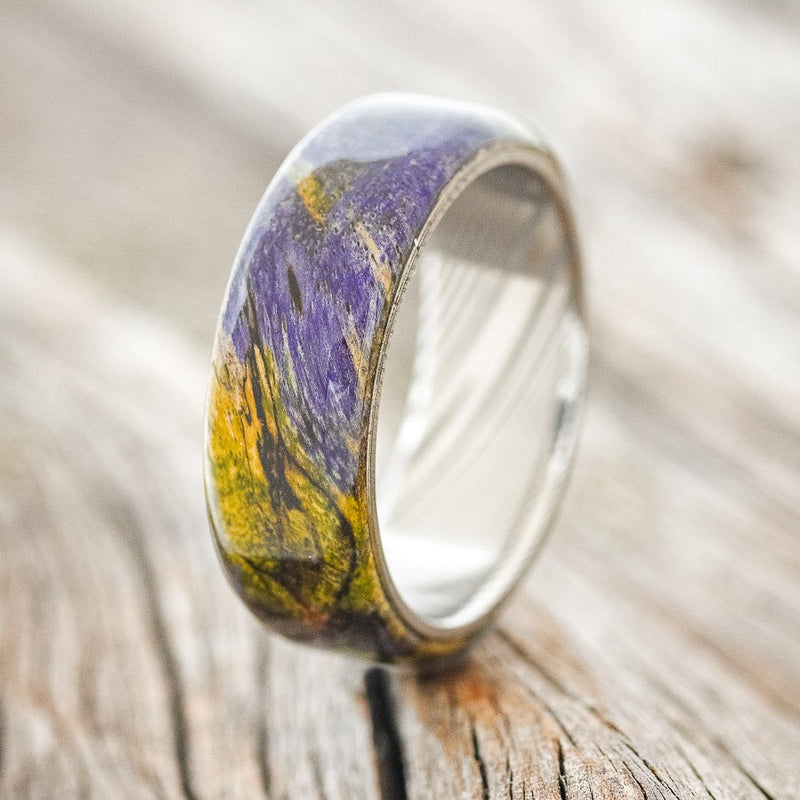 Shown here is "Haven", a custom, handcrafted men's wedding ring featuring a unique green and purple dyed spalted maple wood overlay on a Damascus steel band, upright facing left. This ring was inspired by the Joker character. Additional overlay options are available upon request.