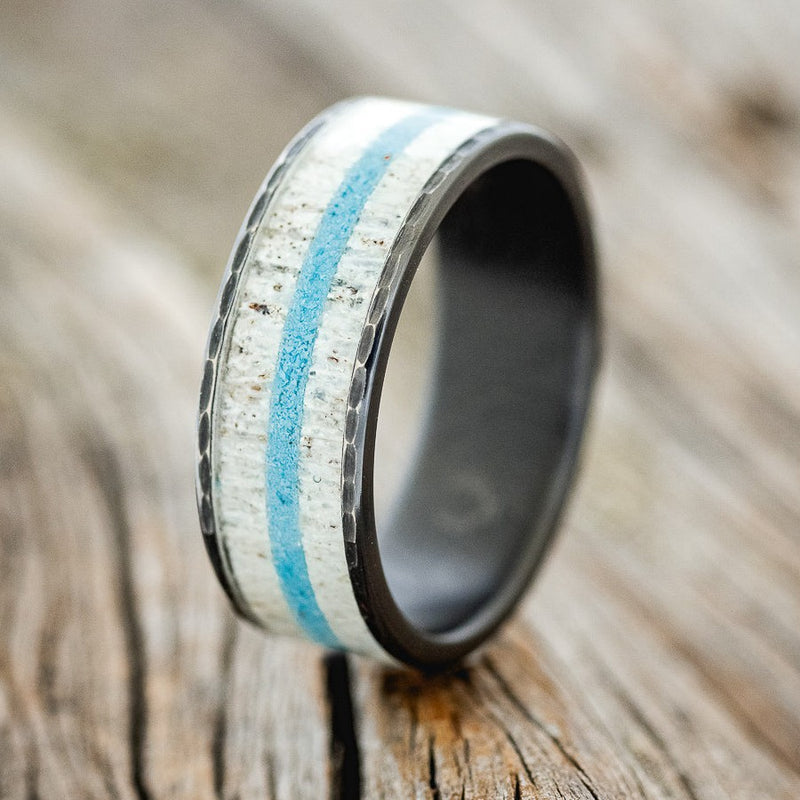 Shown here is "Rainier", a custom, handcrafted men's wedding ring featuring an antler and turquoise inlay on a hammered, fire-treated black zirconium band, upright facing left. Additional inlay options are available upon request.