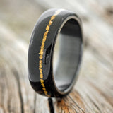 Shown here is "Remmy", a custom, handcrafted men's wedding ring featuring an African black wood overlay and gold nugget inlay, shown here on a fire-treated black zirconium band, upright facing left. Additional inlay options are available upon request.