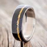 Shown here is "Remmy", a custom, handcrafted men's wedding ring featuring an African black wood overlay and gold nugget inlay, shown here on a titanium band, upright facing left. Additional inlay options are available upon request.