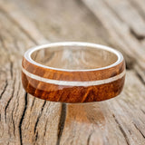 Shown here is "Remmy", a custom, handcrafted men's wedding ring featuring a redwood overlay and an offset mother of pearl inlay, laying flat. Additional inlay options are available upon request.