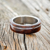 Shown here is "Remmy", a custom, handcrafted men's wedding ring featuring a redwood overlay and an offset mother of pearl inlay on a titanium band, laying flat. Additional inlay options are available upon request.