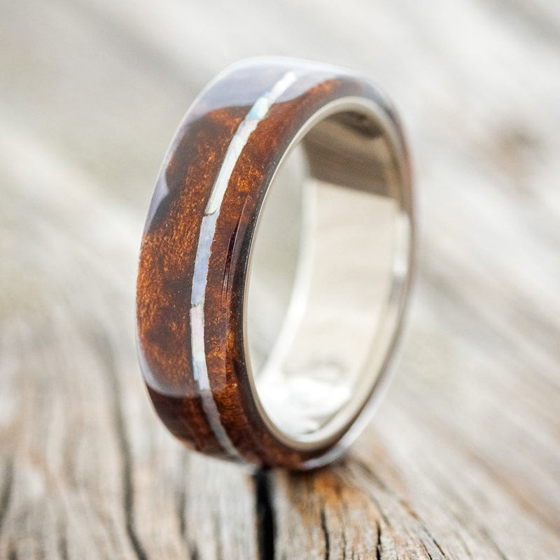 Shown here is "Remmy", a custom, handcrafted men's wedding ring featuring a redwood overlay and an offset mother of pearl inlay on a titanium band, upright facing left. Additional inlay options are available upon request.