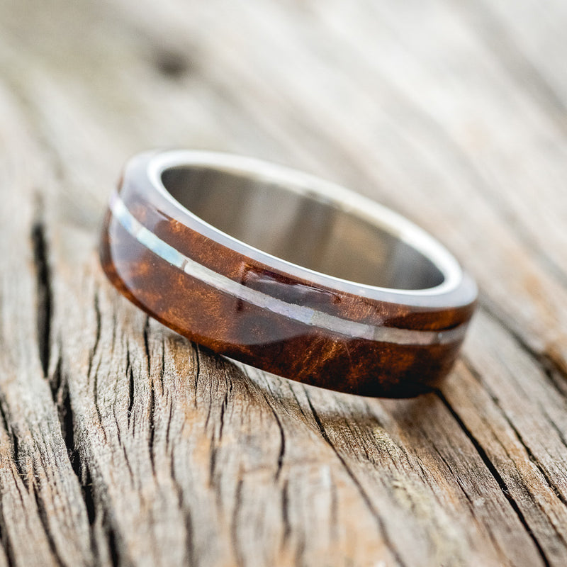 Shown here is "Remmy", a custom, handcrafted men's wedding ring featuring a redwood overlay and an offset mother of pearl inlay on a titanium band, tilted left. Additional inlay options are available upon request.