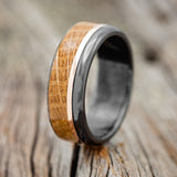 Shown here is "Ezra", a custom, handcrafted men's wedding ring featuring a whiskey barrel oak overlay and a 14K rose gold inlay, shown here on a black zirconium band, upright facing left. Additional inlay options are available upon request.