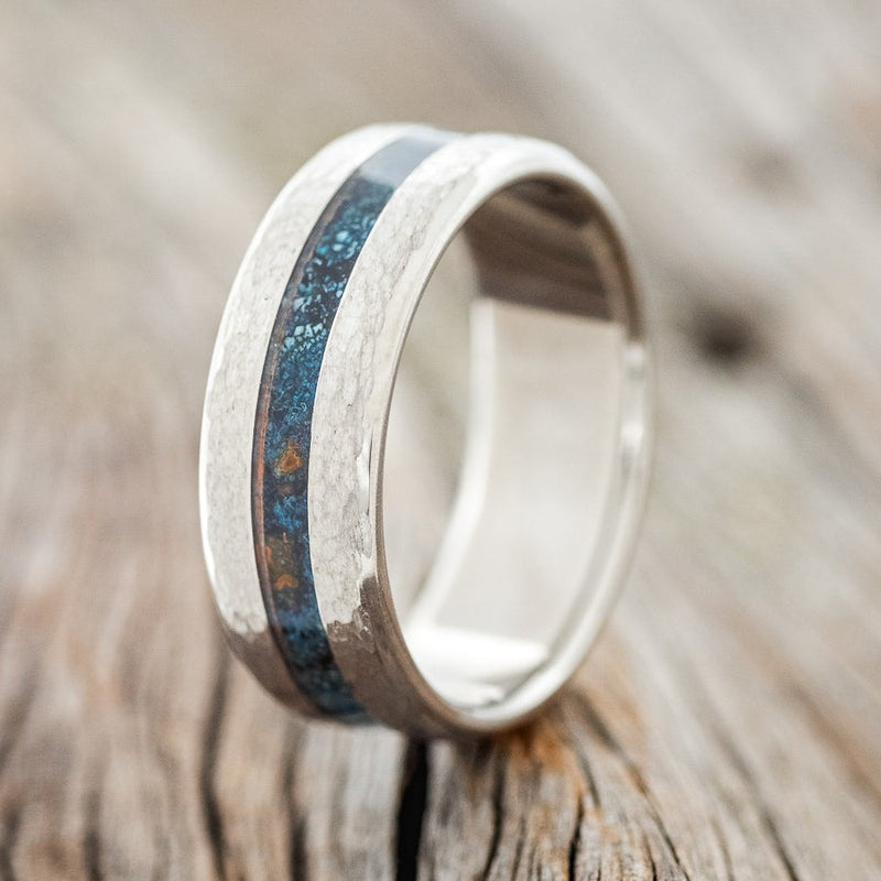 Shown here is "Nirvana", a custom, handcrafted men's wedding ring featuring a centered patina copper inlay and a hammered finish, upright facing left. Additional inlay options are available upon request.