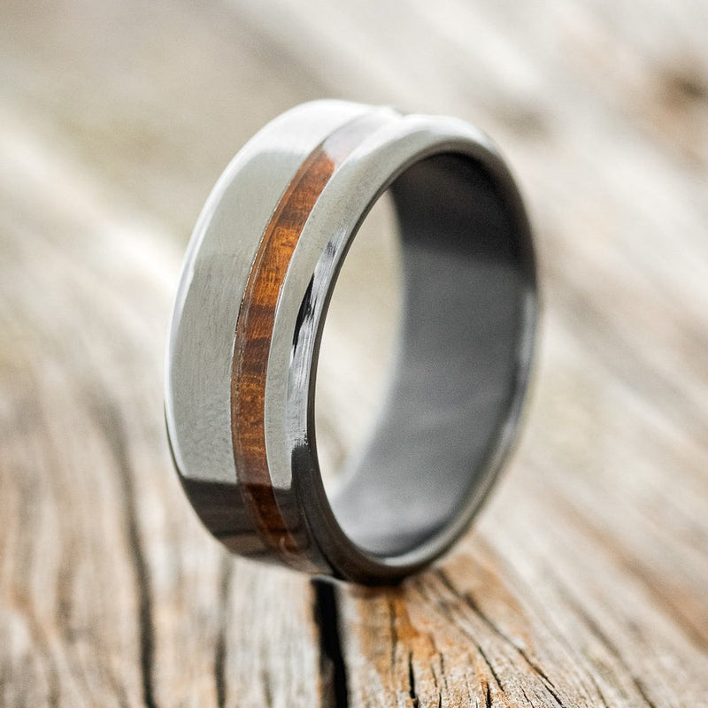 Shown here is "Vertigo", a custom, handcrafted men's wedding ring featuring an offset ironwood inlay on a fire-treated black zirconium band, upright facing left. Additional inlay options are available upon request.