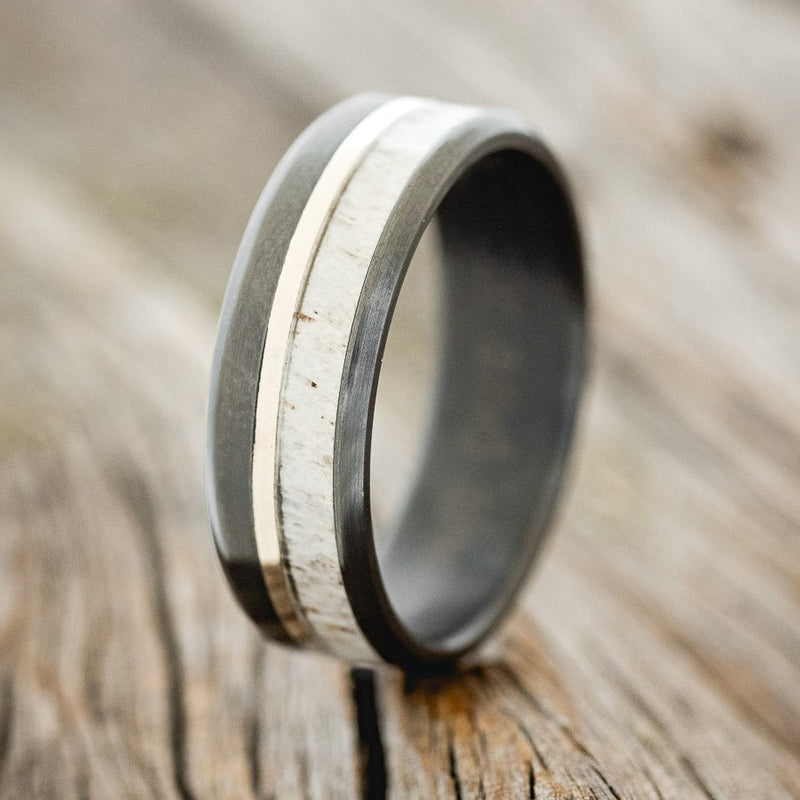 Shown here is "Tanner", a custom, handcrafted men's wedding ring featuring a naturally shed elk antler overlay and a 14K white gold inlay, shown here on a fire-treated black zirconium band, upright facing left. Additional inlay options are available upon request.